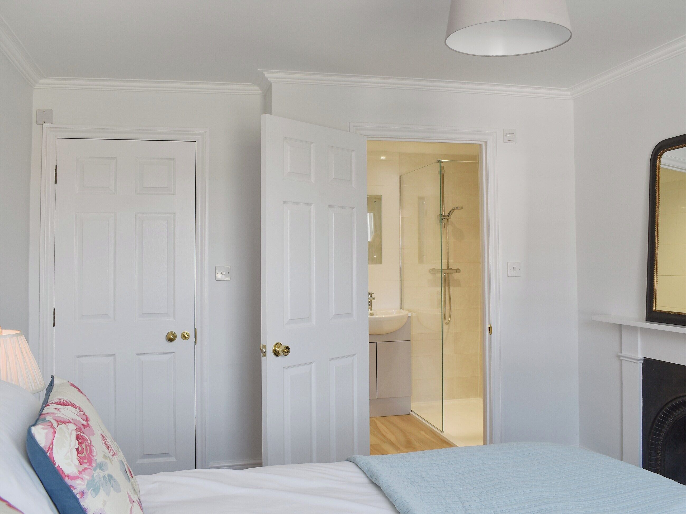 A bedroom with a bed, a nightstand, a lamp, a mirror, and a door leading to a bathroom with a shower.