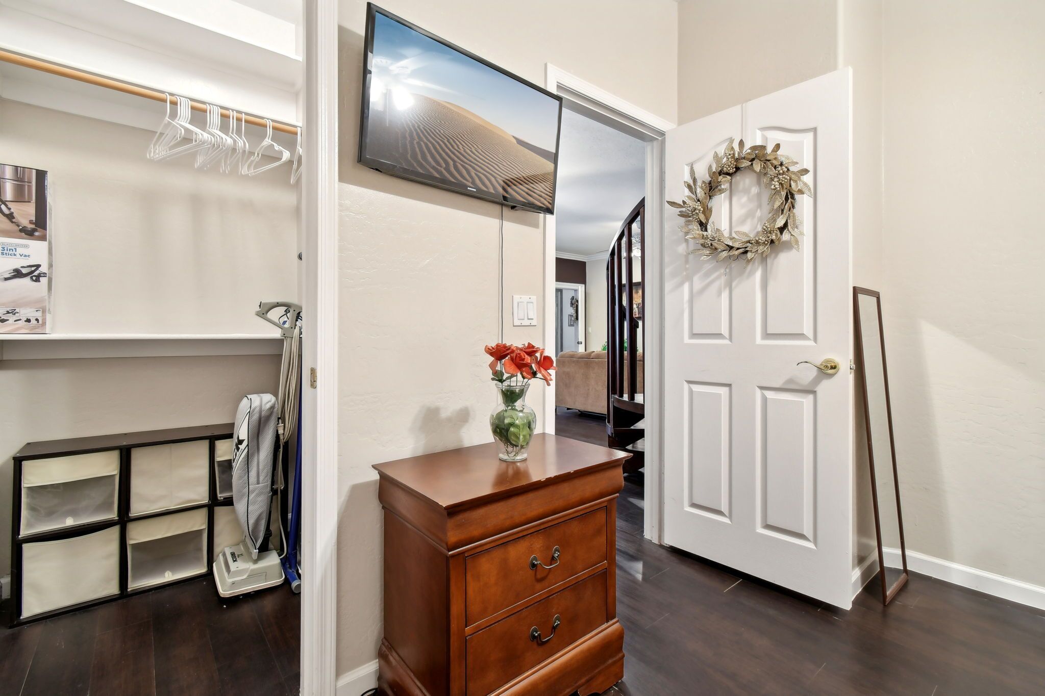 Un couloir avec une commode en bois, un téléviseur fixé au mur, une porte décorée d’une guirlande et un porte-chaussures.