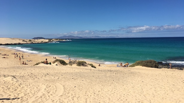 Una playa con aguas turquesas cristalinas, dunas arenosas y algunas personas disfrutando del paisaje.