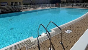 Una piscina azul cristalina con una escalera metálica, rodeada por una valla blanca, y un cielo despejado arriba.