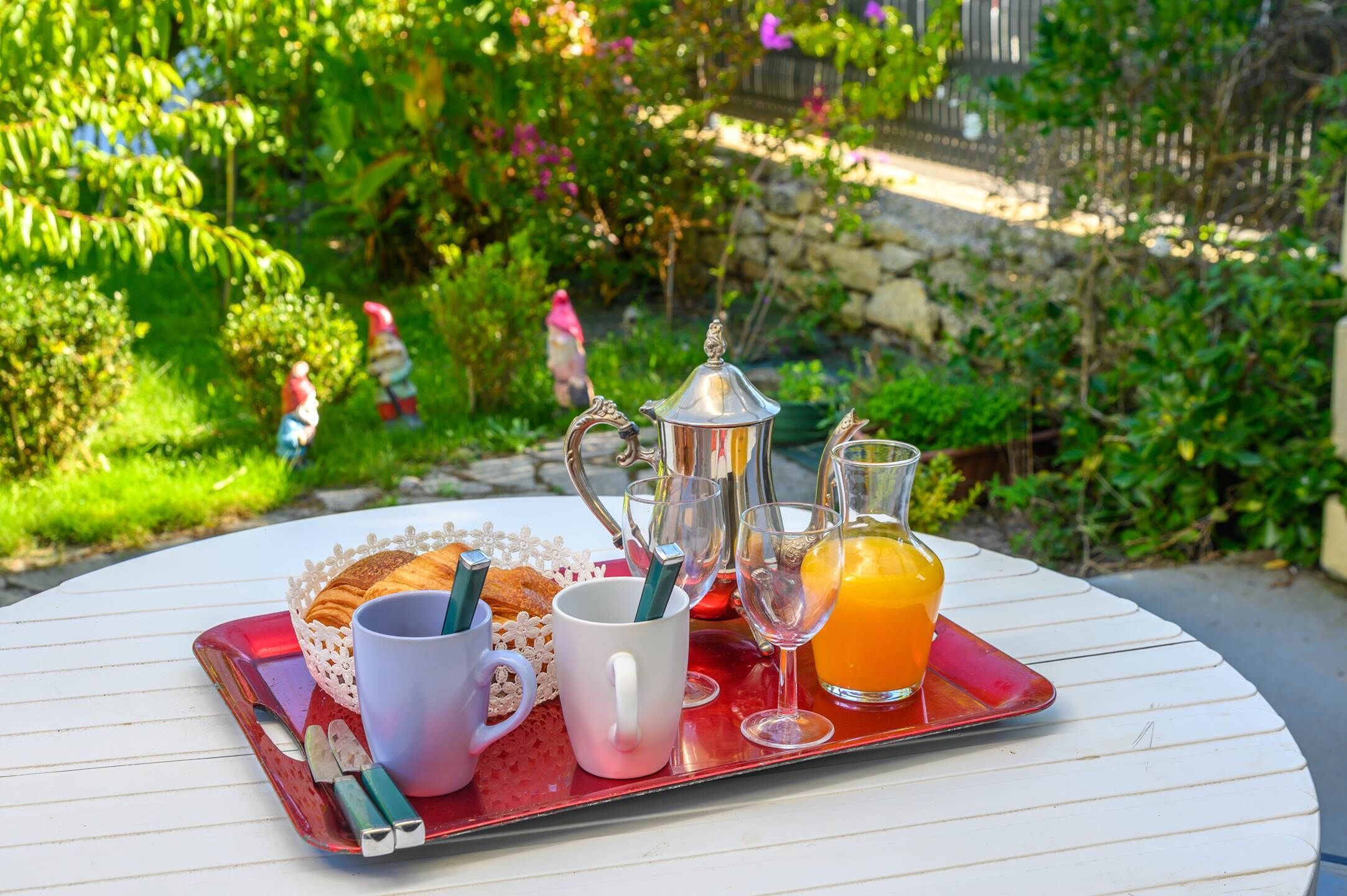Un vassoio colazione con croissant, un teiera, due tazze e un bicchiere di succo d'arancia su un tavolo bianco all'aperto.