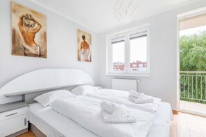 Ein Schlafzimmer mit einem großen Bett, zwei Bildern an der Wand, einem Fenster mit Blick auf Gebäude und einem Balkon mit Grünpflanzen.