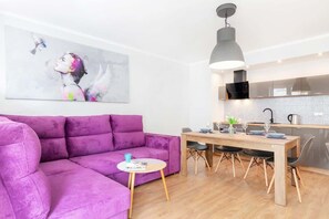 Ein modernes Wohnzimmer mit einem violetten Sofa, einem Esstisch mit Stühlen, einem kleinen runden Tisch mit einer Tasse und einer Küchenzeile mit einer Pendelleuchte.