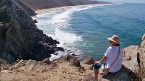 Une personne est assise sur un promontoire rocheux, d’où elle a une vue sur une plage. On peut voir les vagues se briser sur le rivage et des montagnes en arrière-plan.