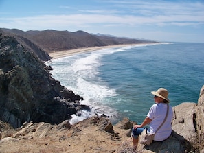 A person sitting on a rocky outcrop overlooking a beach with waves crashing against the shore and mountains in the background.