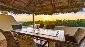 Un espace repas avec une table et des chaises, un toit de chaume et une vue sur le coucher de soleil sur l’océan.