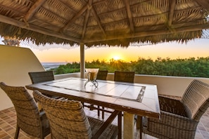 A dining area with a table and chairs, a thatched roof, and a view of the sunset over the ocean.