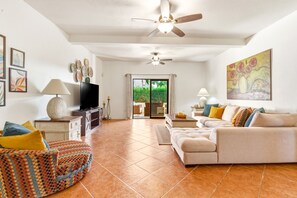 Un salon comprenant un canapé, un fauteuil, un téléviseur, un ventilateur de plafond, des œuvres d’art et une vue sur l’extérieur, visible à travers une porte vitrée.