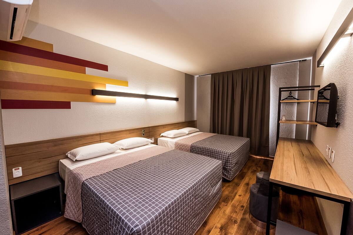 A hotel room with two beds, a wooden desk, a mirror, and a wall-mounted light fixture.