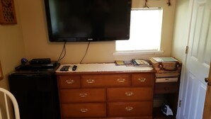 A room with a flat-screen TV mounted on the wall, a wooden dresser with multiple drawers, a small desk with a basket, and a window with blinds.