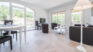 A spacious living area with a dining table, chairs, a sofa, and a TV. Large windows offer views of greenery. - Completely renovated Mas Provencal (Manosque)