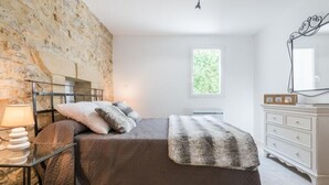 A bedroom with a stone wall, a bed with a grey blanket, a bedside table with a lamp, and a dresser with a mirror. - Completely renovated Mas Provencal (Manosque)