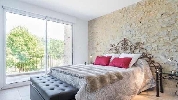 A bedroom with a stone wall, a large window, a bed with a patterned bedspread, and a black bench. - Completely renovated Mas Provencal (Manosque)