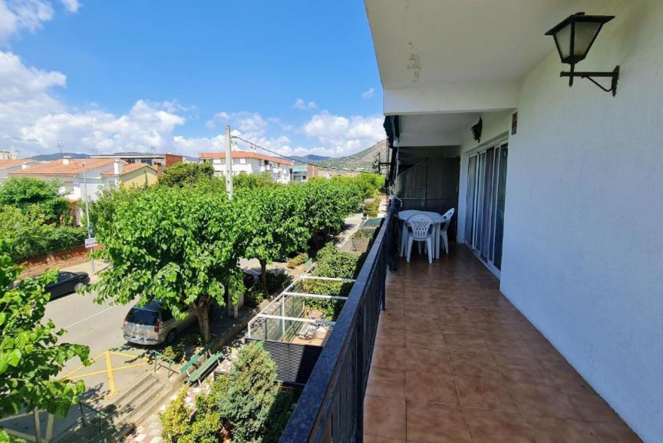 A balcony with a view of a parking area and greenery, a table with chairs, and a lamp.