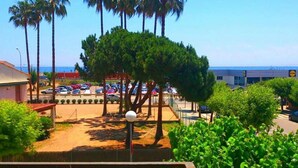 A parking lot with cars, a building with a blue roof, and palm trees. - Apartment Cabrera Burriac (Cabrera de Mar)