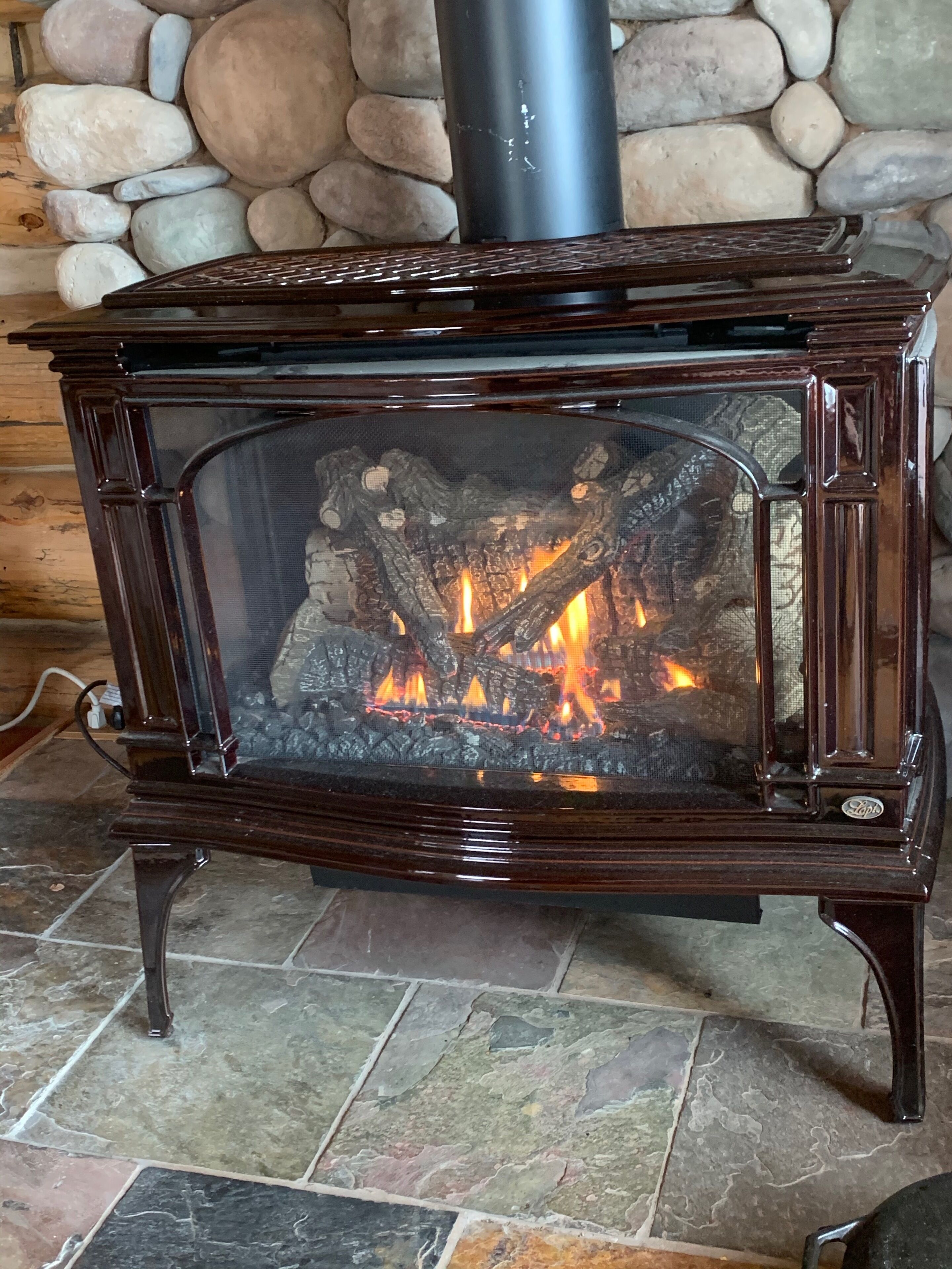 A wood-burning stove with a glass front, set against a stone wall background.