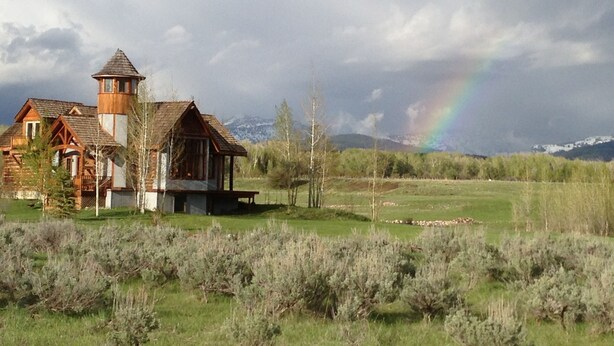 A rustic wooden house with a tower, surrounded by a grassy field and sparse trees.