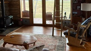 A log cabin interior with a wooden ceiling, a large window, a fireplace, a couch, a coffee table, and a ladder.