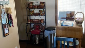 Una cocina con una mesa de madera, un cubo de metal, una repisa con vajilla y un cartel en la pared.