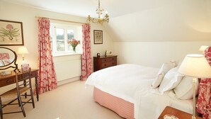 A bedroom with a large bed, a wooden dresser, a desk with a chair, a window with curtains, and a chandelier. - Woodland Cottage, South Moulton, a beautiful, spacious cottage on two floors in a private location (South Molton)