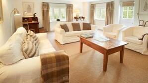 A spacious living room with a white sofa, a wooden coffee table, and two white armchairs. There is a wooden desk with a lamp and a framed picture on the wall. - Woodland Cottage, South Moulton, a beautiful, spacious cottage on two floors in a private location (South Molton)