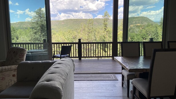 Una habitación con un amplio ventanal que da a una ladera boscosa. Hay un sofá, una silla y una mesa con sillas.