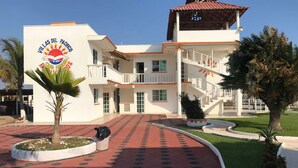 A building with a sign reading "Villas del Pacifico Casa Familia." It features a central courtyard with a red patterned walkway, surrounded by greenery and palm trees. - Villas del Pacifico | Habitación Privada #3 (El Carrizal)