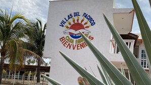 A sign with "Villas del Pacifico" and "Bienvenidos" text, palm trees, and a building in the background. - Villas del Pacifico | Habitación Privada #3 (El Carrizal)