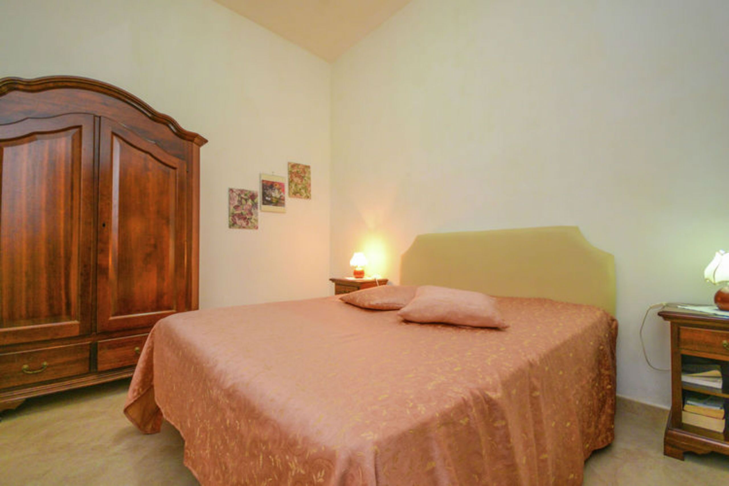 A bedroom with a large wooden wardrobe, a bed with a pink bedspread, a bedside table with a lamp, and a wall-mounted shelf.