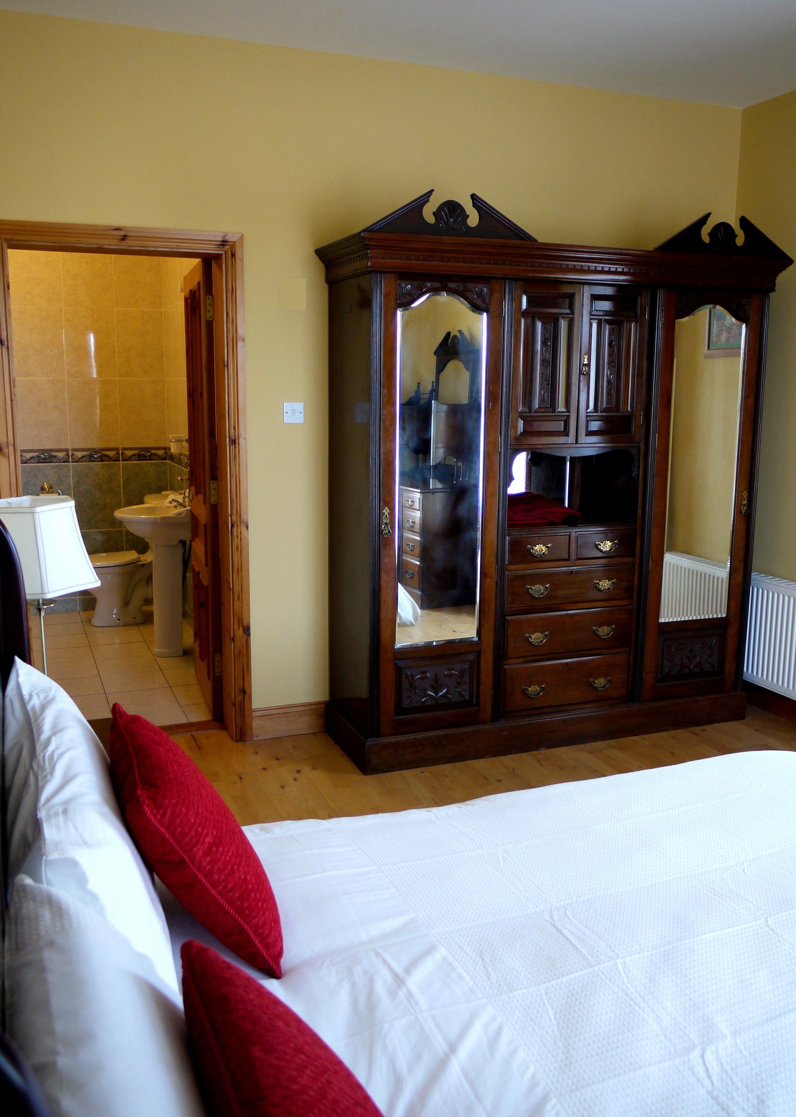 Une chambre à coucher avec une grande armoire en bois, un lit avec des oreillers blancs et rouges, et une salle de bain visible à travers une porte ouverte.