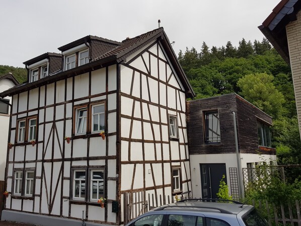 Una casa tradicional con entramado de madera y una extensión moderna, un coche estacionado al frente, y un exuberante fondo verde.