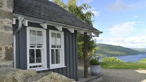 A stone house with a grey exterior and white trim, a covered porch, and a view of a lake and hills in the background. - Beautiful Sky Cottage overlooking Loch Tay (Aberfeldy)