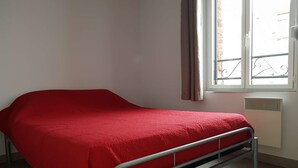 A single bed with a metal frame and a red bedspread in a room with a window and a radiator. - Apartment n°4 (2/4 P.) on 2nd floor without elevator (Berck-sur-mer)