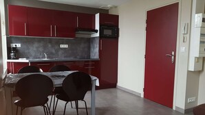 A compact kitchen with red cabinets, a microwave, and a coffee maker. There is a dining table with chairs and a red door. - Apartment n°4 (2/4 P.) on 2nd floor without elevator (Berck-sur-mer)