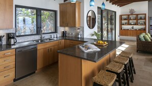 A modern kitchen with wooden cabinets, a central island with stools, a built-in dishwasher, and a stone backsplash. - Eureka Villa! Stunning Views in Paradise! 3BR, 3.5BA (Rendezvous Bay)