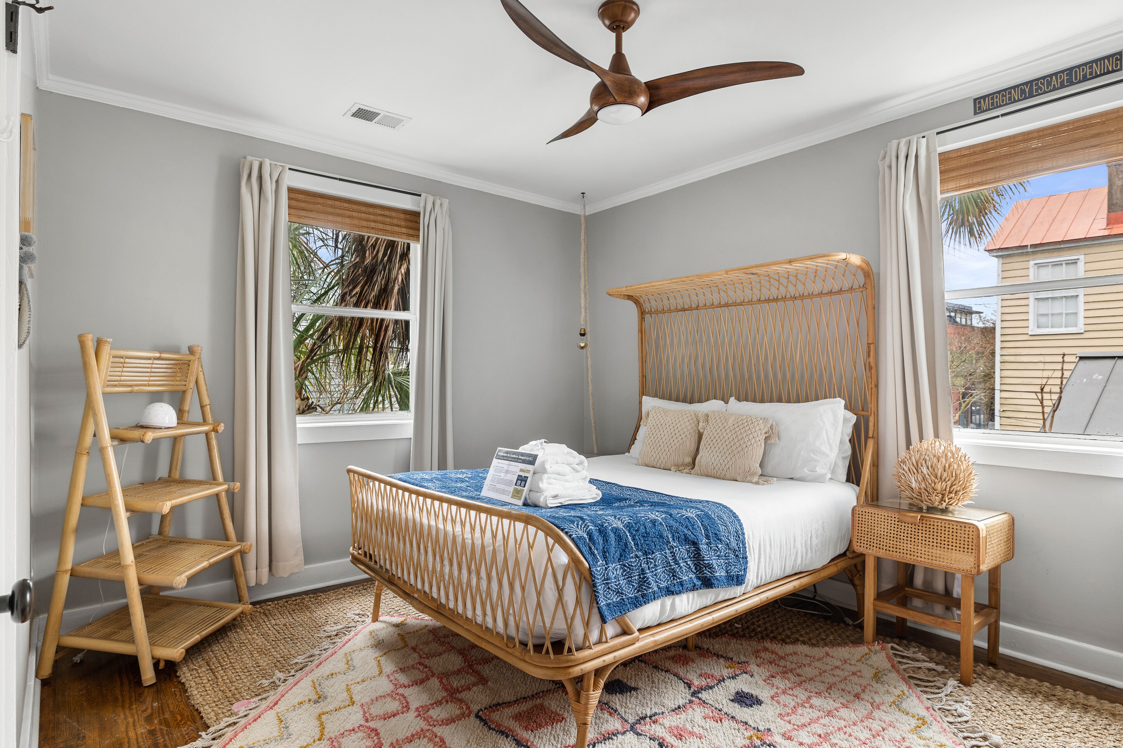 A bedroom with a bed, a ceiling fan, a window with curtains, a wooden chair, and a small table with a decorative item.