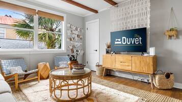 Ein Wohnzimmer mit Fernseher, Holzmöbeln, einem Rattan-Sessel und einem großen Fenster mit Blick auf Palmen.