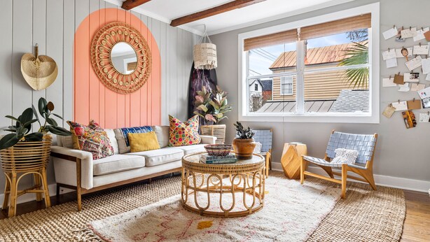 Een gezellige woonkamer met een witte bank, een rond rieten bijzettafeltje en een groot raam met uitzicht op huizen.