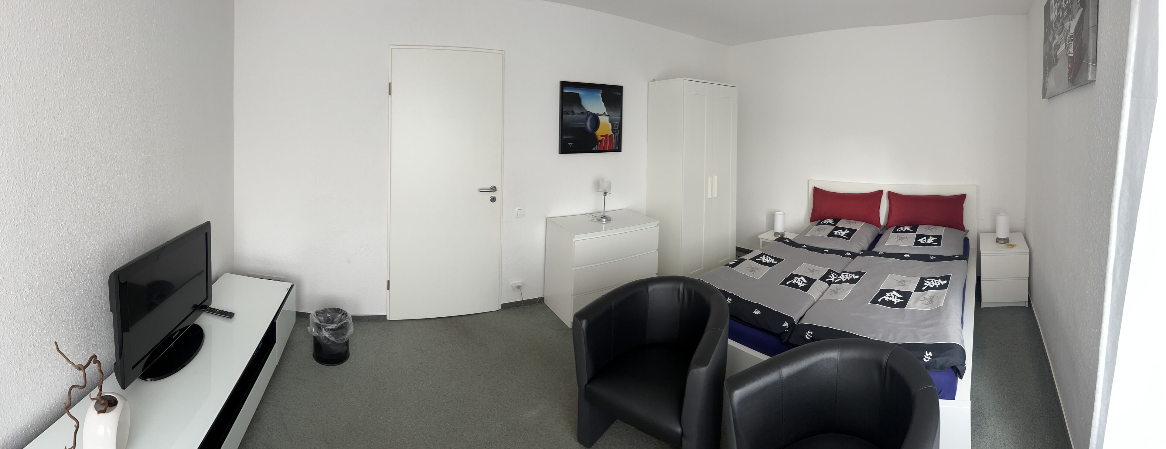 Quarto de hotel compacto com cama, escrivaninha, cadeira, televisão, guarda-roupa e pequena mesa de cabeceira.