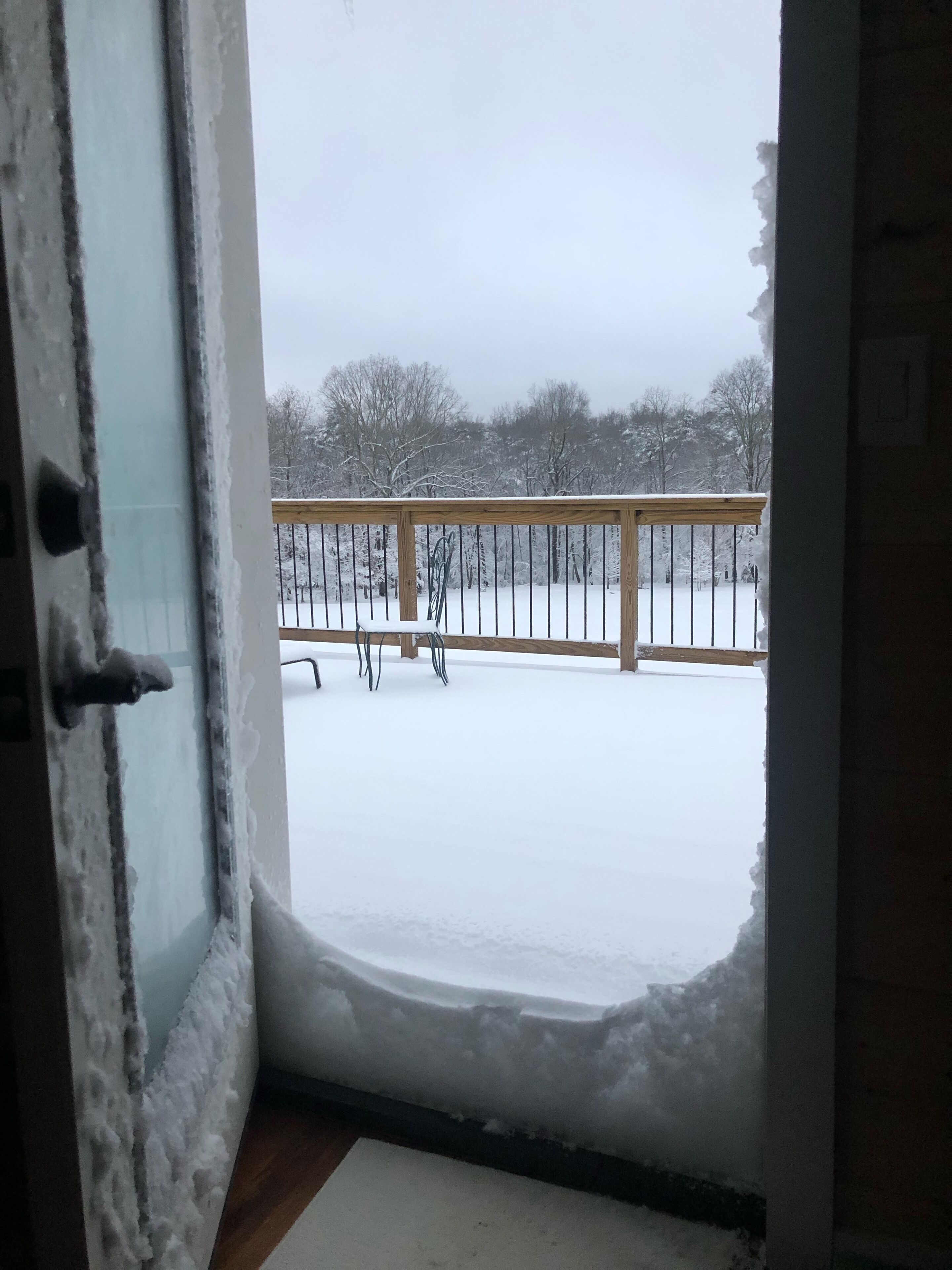 A view from a room with a door leading to a balcony. The balcony has a wooden railing and is covered with snow.
