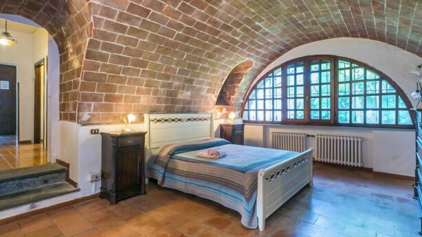 A bedroom with a brick ceiling, a large arched window, a bed, bedside tables, and a radiator. - Poggio al Colle by Interhome (Montaione)