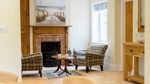 A room with a fireplace, plaid chairs, a table, and a painting of a seascape. - Sea Holly Cottage-UK11931 (Thornham, near Kings Lynn)