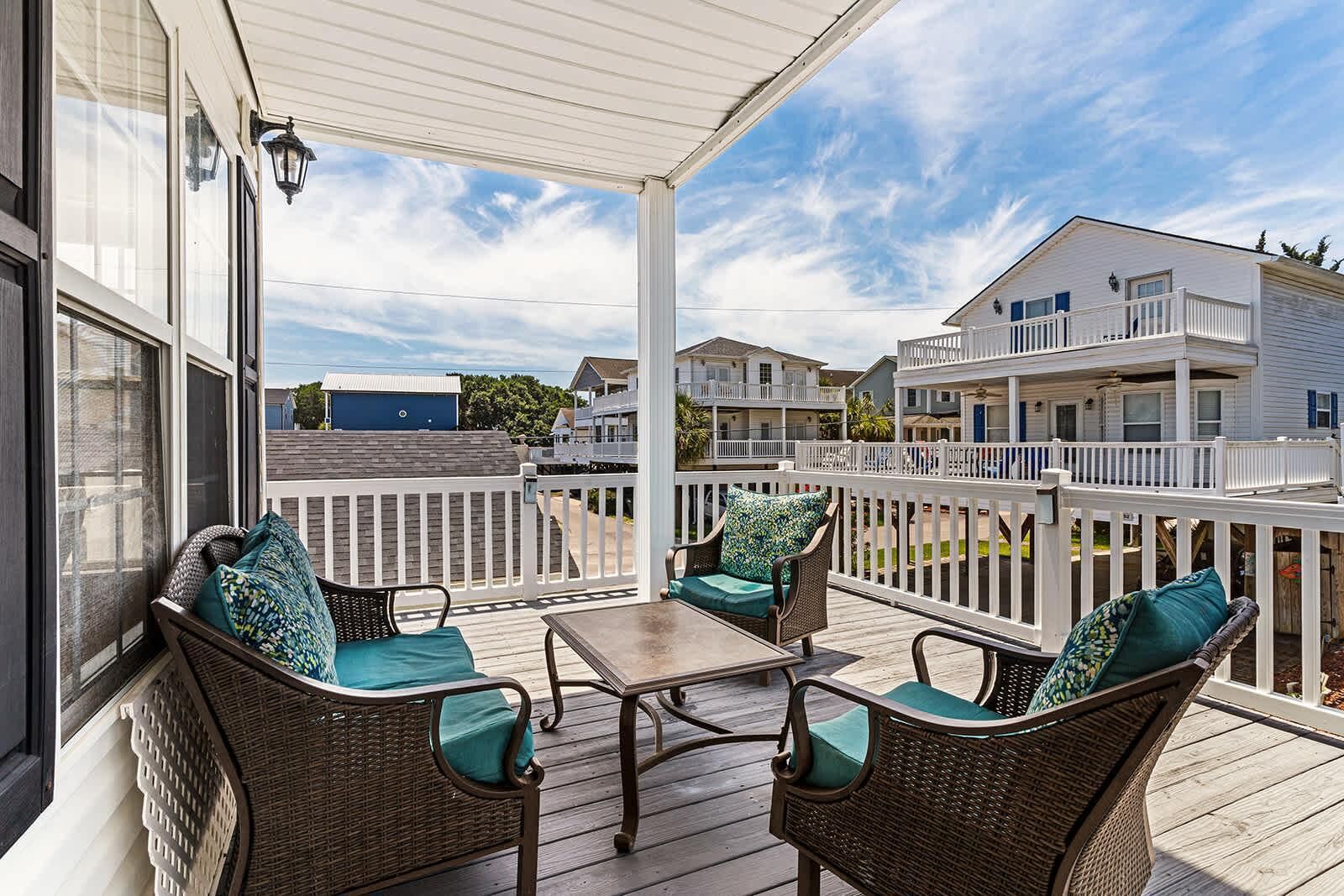 A covered outdoor area with wicker furniture and a table, overlooking a residential area with multiple buildings.