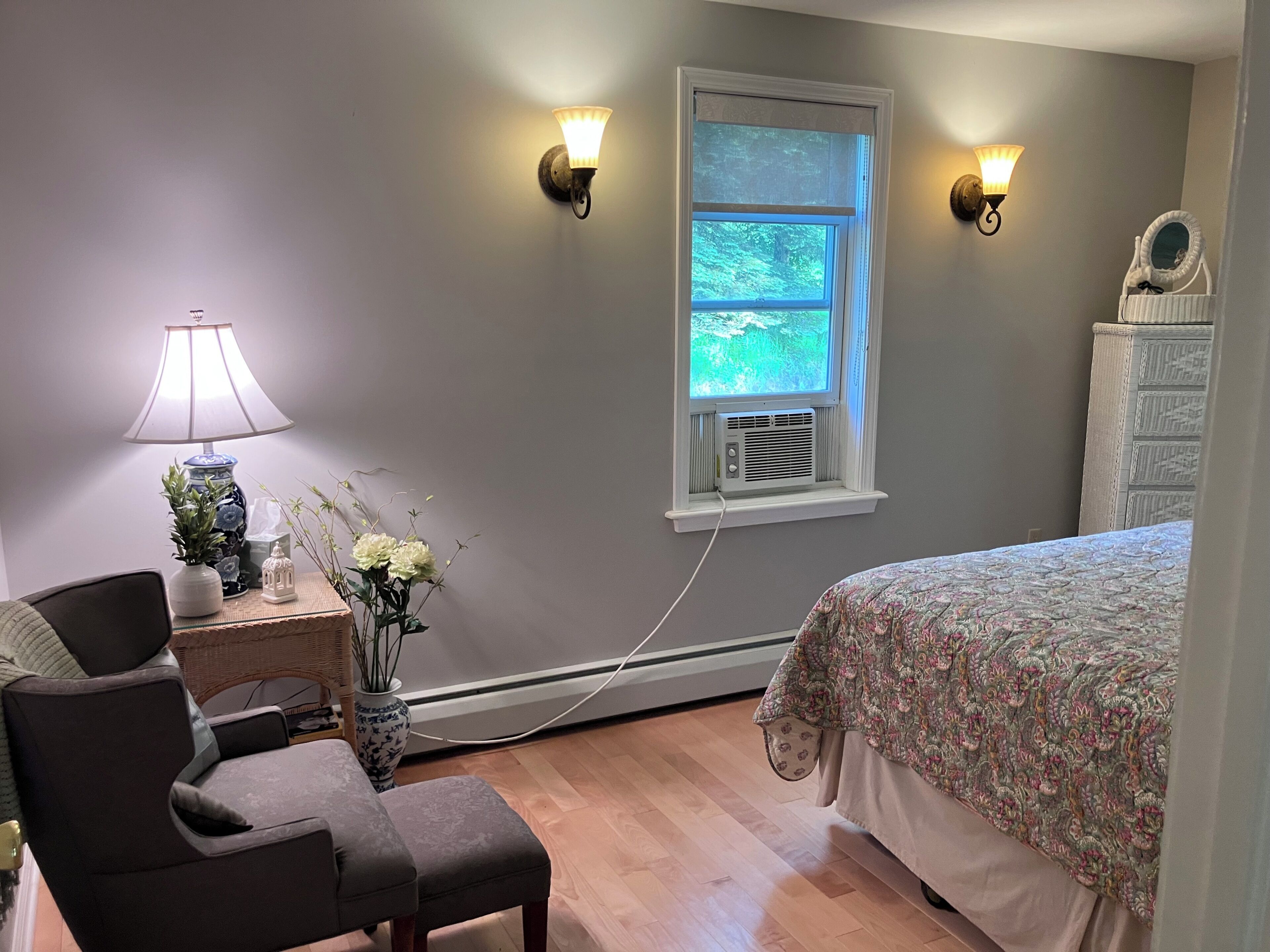 Une chambre à coucher comprenant un lit, une chaise, une lampe, un vase avec des fleurs, une commode et deux appliques murales.