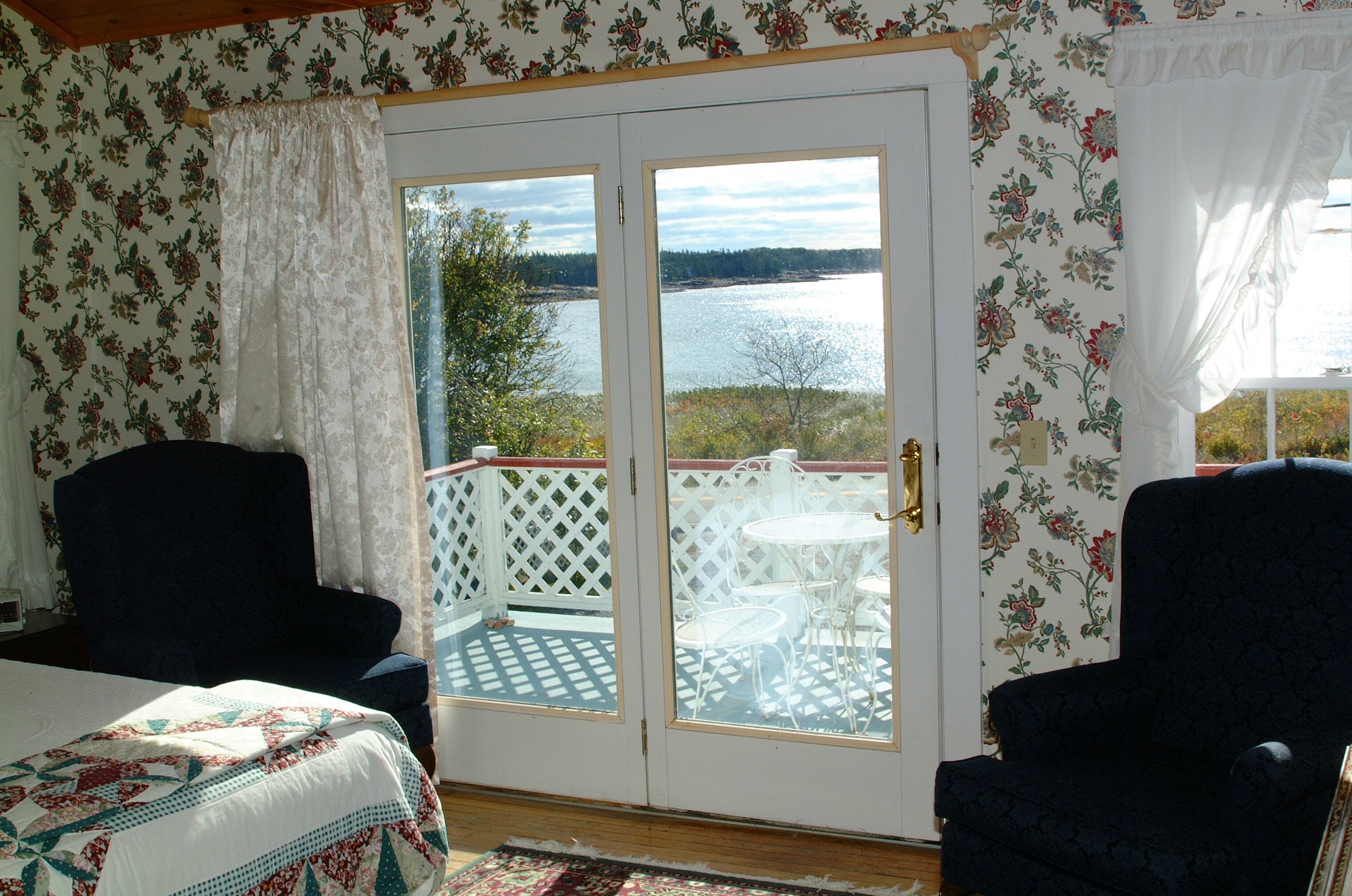 A room with a bed, two chairs, a table, and a view of a lake through a glass door.