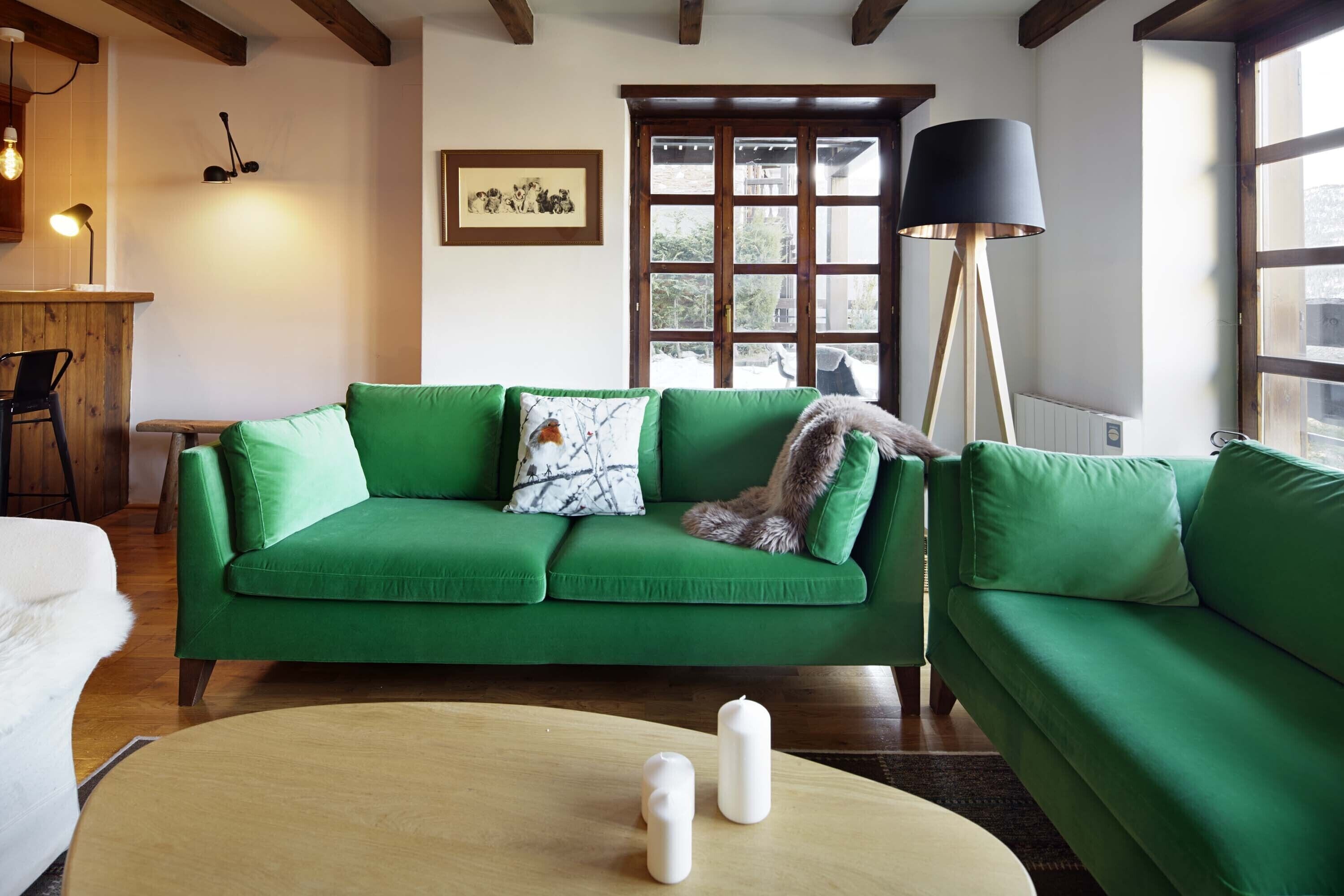 Un sofá verde con un cojín blanco y negro estampado, una mesa de centro de madera con dos velas blancas, una lámpara de pie negra y una ventana con vista a vegetación.