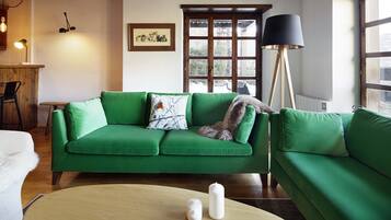 Un sofá verde con un cojín blanco y negro estampado, una mesa de centro de madera con dos velas blancas, una lámpara de pie negra y una ventana con vista a vegetación.