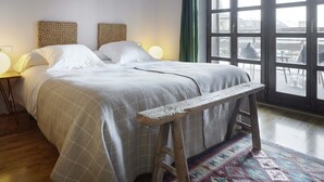 Une chambre à coucher avec un grand lit, deux têtes de lit, un banc, une table de chevet et une fenêtre donnant sur des immeubles.