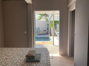A bedroom with a bed, a neatly made bed with folded towels, and a view of an outdoor swimming pool through an open door. - JACARANDA CASE PALM (2 people) (La Saline-Les-Bains)