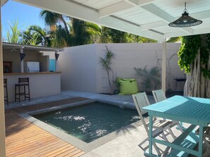 A pool area with a wooden deck, a table with chairs, and a bar area with stools. - JACARANDA CASE PALM (2 people) (La Saline-Les-Bains)